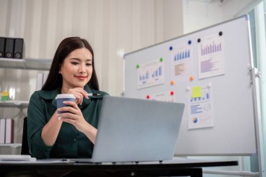 Modern bir ofiste dizüstü bilgisayar üzerinde çalışırken elinde kahve fincanı tutan bir iş kadını. Arkaplanda çizimler ve grafikler var..