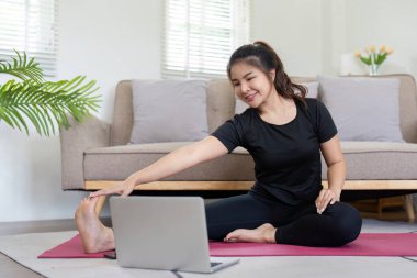 Spor kıyafetli genç bir kadın evde antrenman yaptıktan sonra mindere uzanıyor ve oturma odasında dizüstü bilgisayar izliyor..