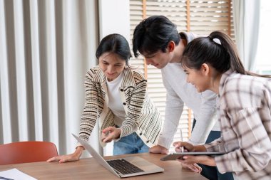 Üç öğrenci, parlak ve modern bir sınıf ortamında dizüstü bilgisayar ve tablet kullanarak bir proje üzerinde birlikte çalışıyorlar..