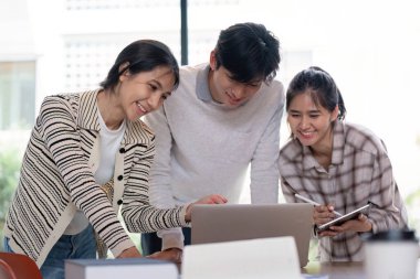 Üç öğrenci bir dizüstü bilgisayarda birlikte çalışıyor. Parlak ve modern bir sınıf ortamında ortak bir proje yürütüyorlar..