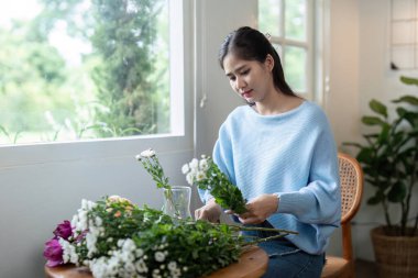 Mavi kazaklı bir kadın, büyük pencereli ve dışarıda yeşil bitkiler bulunan iyi aydınlatılmış bir odada, ahşap bir masada çiçek buketi hazırlıyor..