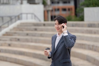 Takım elbiseli, kendine güvenen genç bir işadamı, telefonla konuşuyor, elinde kahve fincanı tutuyor, modern bir şehir ortamında duruyor..