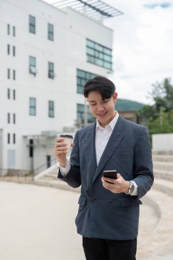 Takım elbiseli genç bir işadamı, güneşli bir günde modern bir ofis binasının önünde akıllı telefonunu kontrol ederken kahve molası veriyor..