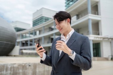 Takım elbiseli genç profesyonel bir adam akıllı telefonunu kullanırken gülümsüyor ve modern bir ofis binasının önünde kahve fincanı tutuyor..