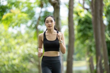 Atletik giysili genç bir kadın yemyeşil bir parkta koşuyor, fitness ve sağlıklı bir yaşam tarzını destekliyor..