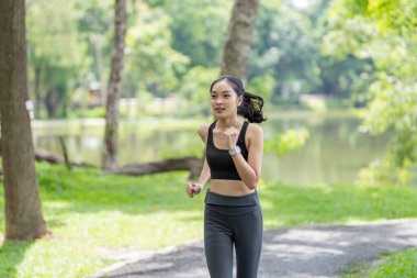 Genç bir kadın parkta koşuyor, spor kıyafetleri giyiyor ve arka planda ağaçlar ve göl var..