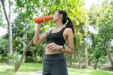 Sporcu genç bir kadın, yeşil bir parkta antrenmanına ara verirken su içiyor..