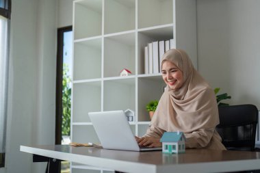 Modern bir ofiste dizüstü bilgisayarla çalışan neşeli, İslami bir emlakçı. Masasında model bir ev var..