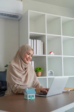 Gülümseyen İslami emlakçı tesettür içinde modern bir ofiste dizüstü bilgisayarda çalışıyor, masada da model bir ev var..