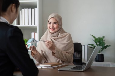 Geleneksel kıyafetli bir Müslüman emlakçı modern ofis ortamındaki bir müşteriyle mülkiyet detaylarını tartışıyor..