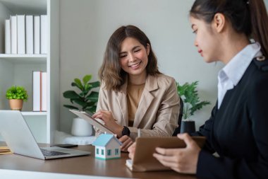 Profesyonel bir emlakçı, modern bir ofisteki bir müşteriyle mülk detaylarını tartışıyor. Her ikisi de belgeler ve bir dizüstü bilgisayarla konuşuyorlar..