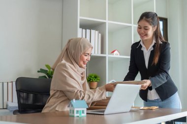 Bir tesettürdeki Müslüman emlakçı modern bir ofiste müşteriyle mülkiyet anlaşmaları tartışıyor, iş dünyasındaki profesyonellik ve kültürel çeşitliliği gösteriyor.