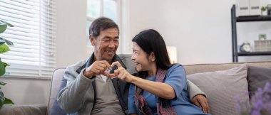 Neşeli yaşlı çift evde birlikte vakit geçiriyorlar, elleriyle kalp şekli oluşturuyorlar, aşkı ve arkadaşlığı simgeliyorlar..