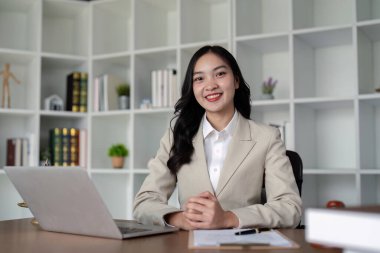 Profesyonel ofis ortamında kendine güvenen bir kadın avukat, arka planda dizüstü bilgisayarı ve kitaplığı olan bir masada oturuyor..