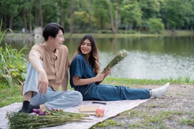 Genç bir çift gölün kenarındaki battaniyeye oturmuş yemyeşil bir parkta huzurlu bir pikniğin tadını çıkarıyorlar..