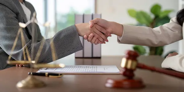 İki avukat, elinde yasal belgeler, adalet terazisi ve bir tokmak olan bir masanın üzerinde el sıkışırken anlaşma ve ortaklığı simgeliyor..