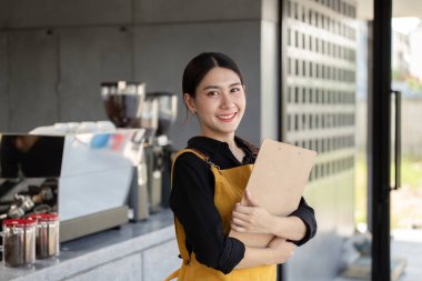 Sarı önlüklü gülümseyen barista. Çağdaş bir kahve dükkanında elinde pano var. Sipariş almaya hazır..