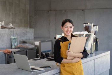 Sarı önlük giyen neşeli bir barista, çağdaş bir kahve dükkanında elinde dizüstü bilgisayarı olan bir pano tutuyor..