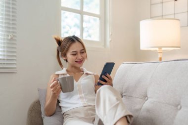 Genç bir kadın, akıllı telefonuyla kahvesini yudumlarken, parlak, modern bir oturma odasında rahat bir şekilde oturuyor..