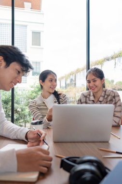 Bir grup öğrenci, geniş pencereli, parlak ve modern bir çalışma alanında bir dizüstü bilgisayar kullanarak bir proje üzerinde birlikte çalışıyorlar..