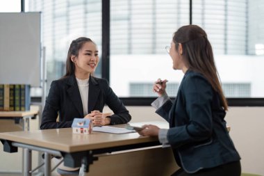 Profesyonel bir emlakçı, modern ofis ortamında bir müşteriyle mülkiyet detaylarını tartışıyor, iş kıyafetleri ve model bir ev sergiliyor..