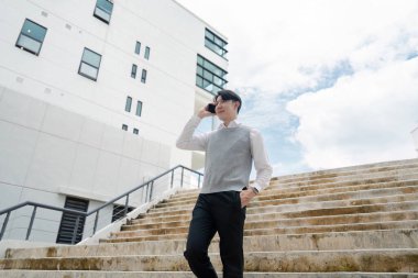 İş kıyafeti giymiş genç bir profesyonel, modern bir ofis binasının önünde, parlak bir gökyüzünün altında cep telefonuyla konuşurken merdivenlerden iniyor..