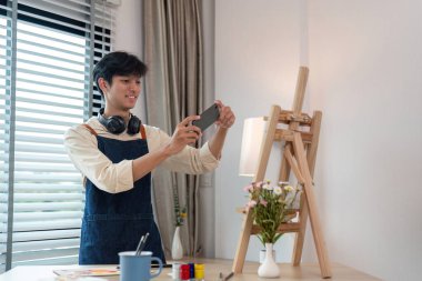 Modern bir stüdyoda, yaratıcı aletler ve doğal ışıkla çevrili akıllı bir telefonla sanat eserinin fotoğrafını çeken genç bir sanatçı..