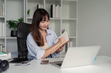 Genç bir öğrenci evde online çalışıyor, dizüstü bilgisayar kullanıyor ve modern, düzenli bir çalışma alanına not alıyor..