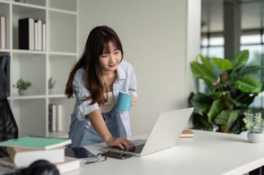 Genç bir öğrenci evde bilgisayar kullanarak ve elinde kahve fincanı tutarak çalışıyor. Modern, parlak bir odada bitkiler ve kitaplarla..