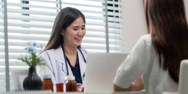 Gülümseyen bir doktor, çağdaş bir muayenehanede bir hastayla görüşür. Masasında ilaç şişeleri bulunur..