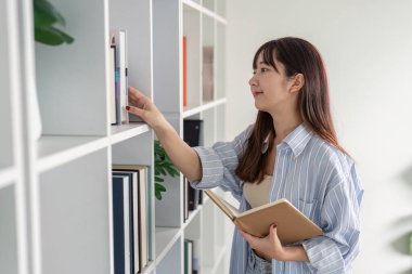 Genç bir öğrenci evde online olarak okuyor, raftaki bir kitaba uzanıyor, modern eğitimi ve uzaktan eğitimi simgeliyor..