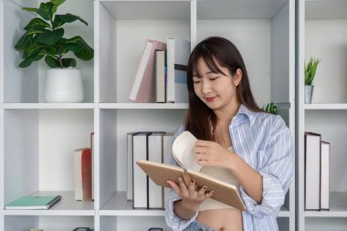 Evde online okuyan genç bir öğrenci, kitaplarla çevrili ve rafları ve bitkileri olan modern bir çalışma alanında bir dizüstü bilgisayar..