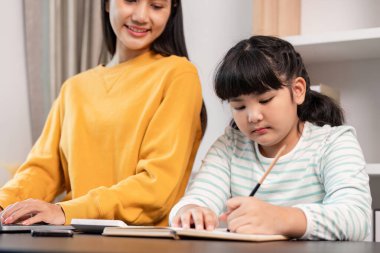 Bir anne ve kızı evde birlikte vakit geçiriyorlar. Genç kız deftere yazmaya odaklanırken anne arka planda sıcak bir şekilde gülümsüyor..