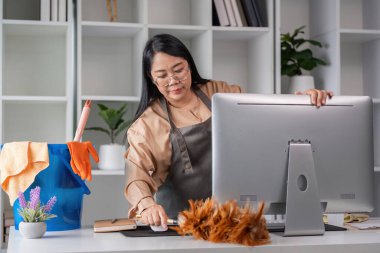 Modern bir ofiste masa ve bilgisayar temizleyen bir kadın. Etrafı temizlik malzemeleri ve bitkilerle çevrili..