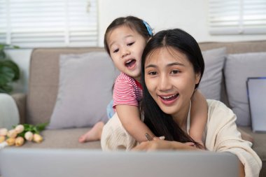 Neşeli bir anne ve küçük kızı rahat bir evde, kanepede dizüstü bilgisayar kullanırken mutlu bir anı paylaşıyorlar..