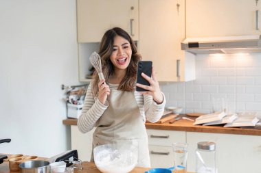 Gülümseyen kadın modern mutfağında akıllı telefonuyla çevrimiçi pişirme dersi alıyor. Mutfak becerileri ve teknolojik öğrenme için mükemmel..