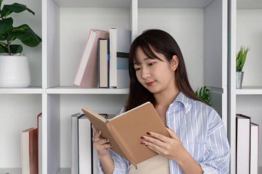 Genç bir kadın, geleneksel öğrenme ve kişisel gelişimin önemini yansıtan ev kütüphanesinde bir kitap okumaya daldı..