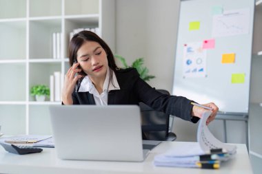 Modern bir ofiste birden fazla işi olan profesyonel bir kadın, iş amaçlarına ulaşmak için telefon görüşmeleri, evrak işleri ve dizüstü bilgisayarları birleştiriyor..