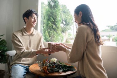 Parlak bir pencerenin önünde gülümseyen bir çift, sıcaklığı ve bağlantıyı vurgulayarak minnetle hediyelerini veriyor..