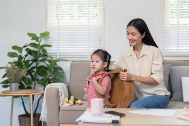 Asyalı anne kızını okula hazırlıyor, etrafı oyuncaklarla çevrili oturma odasında..