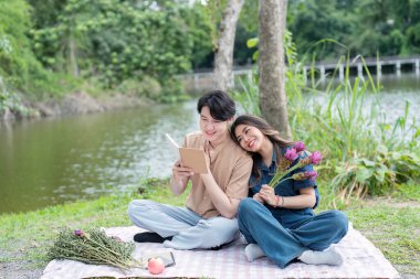 Asyalı bir çift gölün kenarında oturuyor, kitaplar ve çiçeklerle romantik bir piknik yapıyorlar, huzurlu bir ortam yaratıyorlar..