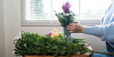 Asyalı kadın bir vazoda renkli bir çiçek düzenleme hazırlıyor, parlak ışıklı bir odada bir masada oturuyor..
