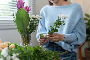 Asyalı kadınların elleri masanın üstündeki vazoda çiçekleri incelikle düzenlerken, iç mekandaki huzur ortamını yansıtıyor..