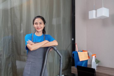 Modern bir ev ortamında elektrikli süpürge tutarken güvenle gülümseyen Asyalı bir kadın kahya. Temizlik görevlerine hazır..