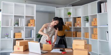 İki kadın e-ticaret işlemleri için paketler hazırlıyor, bant kullanıyor ve iyi donanımlı bir ofis alanında kutular düzenliyor..