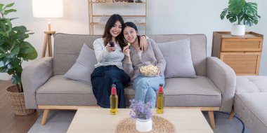 Atıştırmalıklarla televizyon izleyen mutlu bir lezbiyen çift, sıcak ve davetkar bir oturma odasında birbirlerine eşlik ediyorlar..