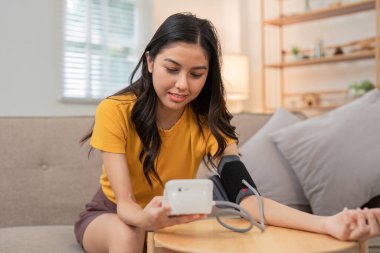 Dijital tansiyon monitörü kullanarak koltuğunda oturan genç bir kadın sağlığını ve sağlığını korumaya odaklanıyor..