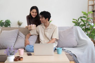 Genç bir çift, rahat bir oturma odasında, dizüstü bilgisayarı ve hediye kutusuyla, bitkilerle çevrili ve rahat bir dekorasyonla birlikte vakit geçiriyorlar..