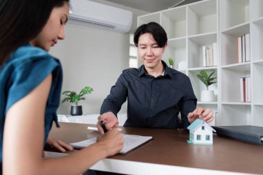 Bir emlakçı ve bir müşteri, başarılı bir alışverişi sembolize eden, üzerinde kağıtlar ve masanda bir model ev bulunan bir evin satışını sonuçlandırdılar..