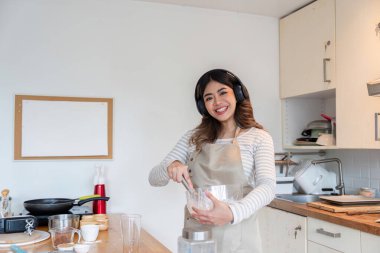 Neşeli bir kadın kulaklık takarken malzemeleri karıştırıyor, hem müzikten hem de ev yapımı pişirmeden zevk alıyor..
